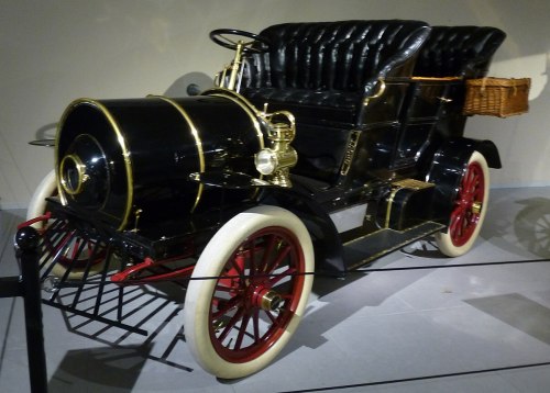 A vintage black car with a shiny finish, red wheels, and a wicker basket, showcasing classic early 20th-century design.