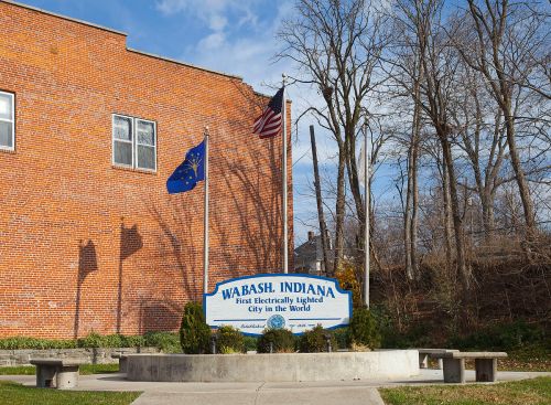 Sign for Wabash, Indiana, noting it as the first electrically lit city in the world, with flags and a brick building in the background.