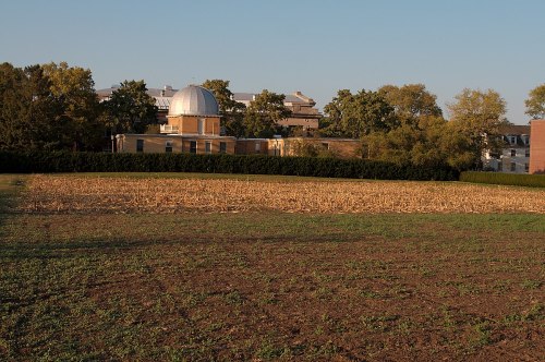 A field with harvested corn stubble in the foreground, and a building with a dome in the background, surrounded by trees.
