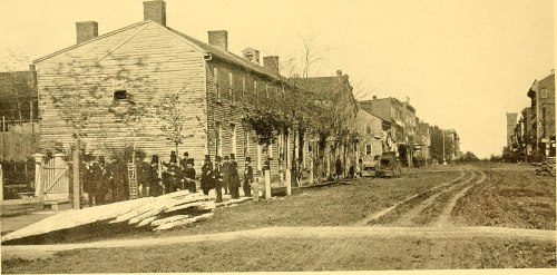 Historic street scene featuring wooden buildings and a group of people, with dirt roads and trees lining the area.