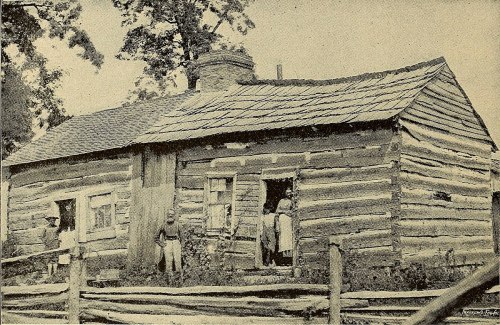 A rustic log cabin with a thatched roof, surrounded by trees, featuring people standing in doorways.