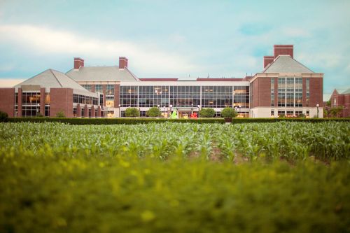 A large brick building with glass windows, surrounded by green fields under a cloudy sky.