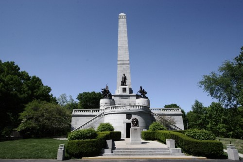 A tall stone monument with statues, surrounded by greenery and a clear blue sky.
