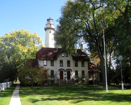 A historic lighthouse beside a large white building, surrounded by trees and a well-maintained lawn.