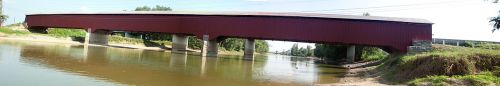 A red covered bridge spans a calm river, supported by concrete pillars, surrounded by greenery and blue sky.