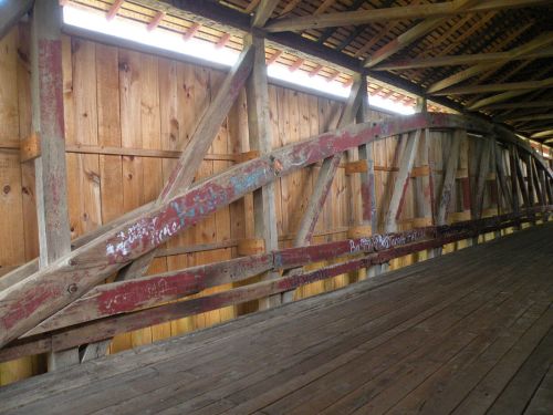Interior view of a wooden covered bridge, featuring rustic beams and a wooden floor with some graffiti on the walls.