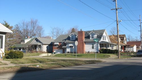 A quiet residential street with various houses, trees, and utility poles under a clear blue sky.