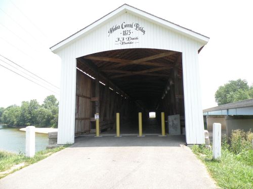 A covered bridge with a white exterior and yellow posts, leading to a river, under a clear sky.