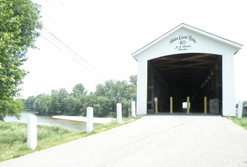 A covered bridge with a white exterior, leading to a river and green trees, under a bright sky.