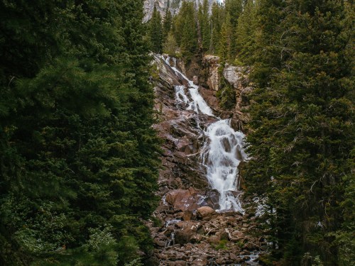 A cascading waterfall flows down rocky terrain, surrounded by lush green trees and a mountainous backdrop.