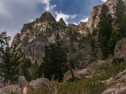Majestic mountain peaks rise above a rocky landscape, surrounded by lush green trees under a cloudy sky.