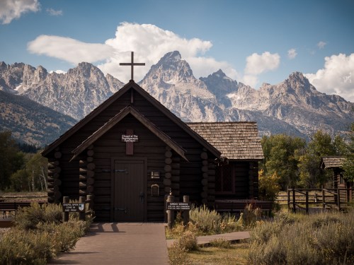 A rustic log chapel with a cross, set against a backdrop of majestic mountains and blue skies.