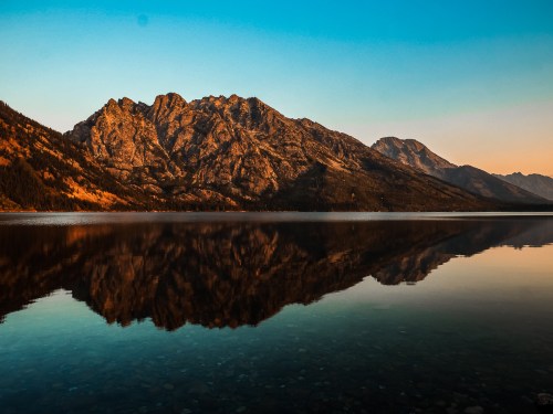 A serene lake reflects majestic mountains under a clear blue sky at sunset.