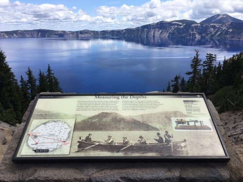 A scenic view of Crater Lake with a historical information sign about measuring its depths in the foreground.