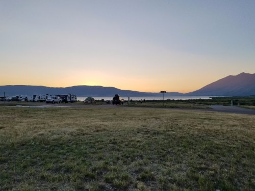A serene sunset over a lake, with camping tents and RVs in the foreground and mountains in the background.