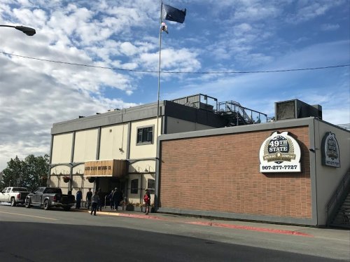Exterior of the 49th State Brewing Co. building, featuring a flag, sign, and people outside on a cloudy day.