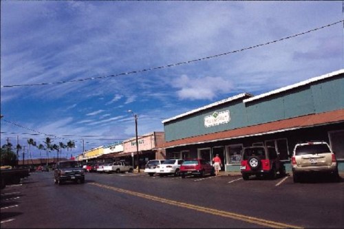 A street view of a shopping area with colorful storefronts, parked cars, and a blue sky with scattered clouds.