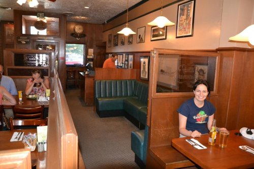 A cozy restaurant interior with wooden booths, a smiling woman at a table, and patrons in the background.