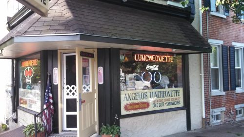 A cozy luncheonette with a sign displaying "Ángelo's Luncheonette" and a menu of sandwiches and cold cuts.