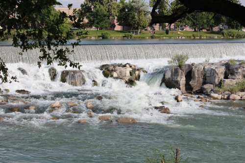 A serene river scene with cascading water over rocks, surrounded by greenery and a park in the background.