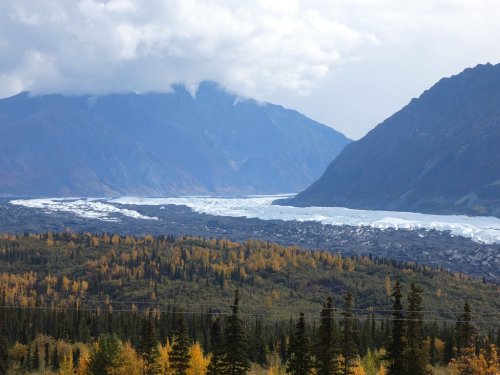 A scenic view of a glacier flowing between mountains, surrounded by autumn foliage and cloudy skies.