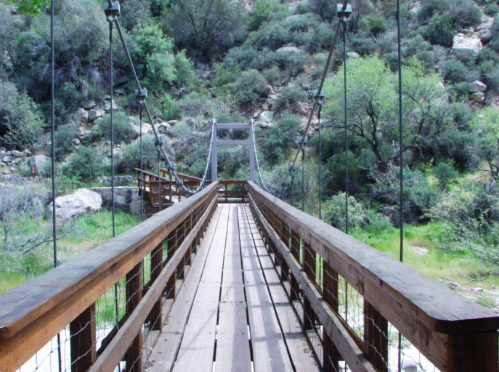 A wooden suspension bridge stretches over a lush, green landscape with trees and rocky terrain in the background.
