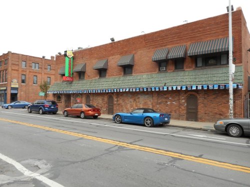 A brick building with a green awning and colorful signage, parked cars lined along the street in front.