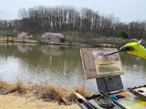 An artist paints a serene landscape by a lake, with blooming trees in the background and a palette of colors nearby.