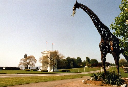 A large metal giraffe sculpture stands in a park, with a lighthouse and a white building in the background.