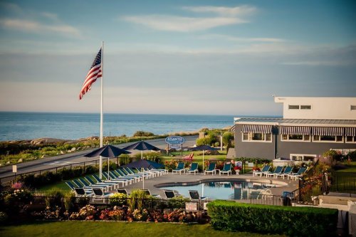 A serene seaside view featuring a pool, lounge chairs, and an American flag, with the ocean in the background.