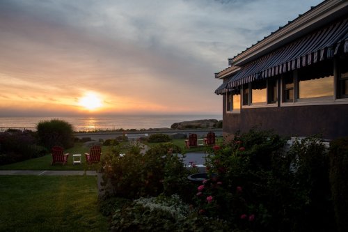 Sunset over the ocean viewed from a garden with chairs, featuring a house with striped awnings in the foreground.