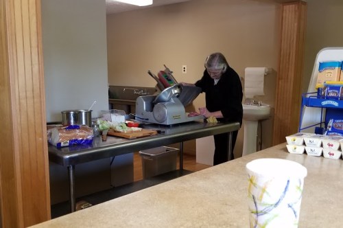 A person using a meat slicer at a kitchen counter, with food items and utensils nearby.