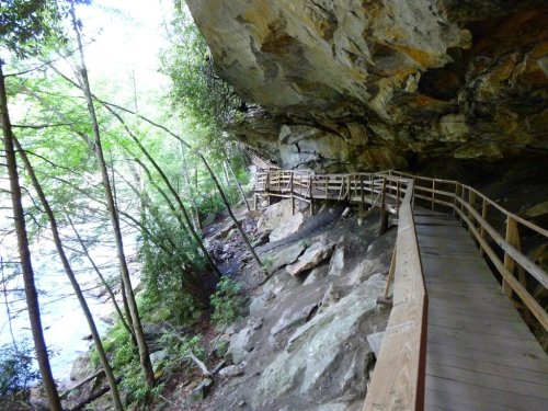 A wooden walkway curves along a rocky riverbank, surrounded by lush greenery and a large overhanging cliff.