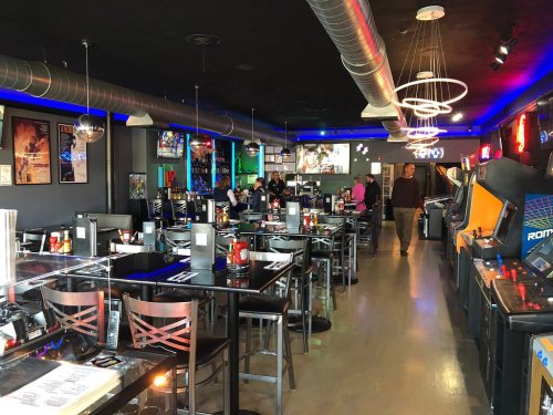 A modern restaurant with arcade games, black tables, and colorful lighting, featuring patrons enjoying their meals.
