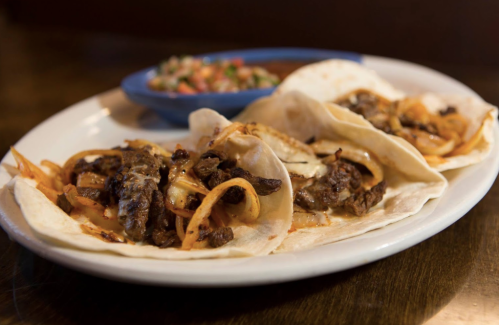 Three tacos filled with grilled meat and onions, served with a side of pico de gallo on a wooden table.