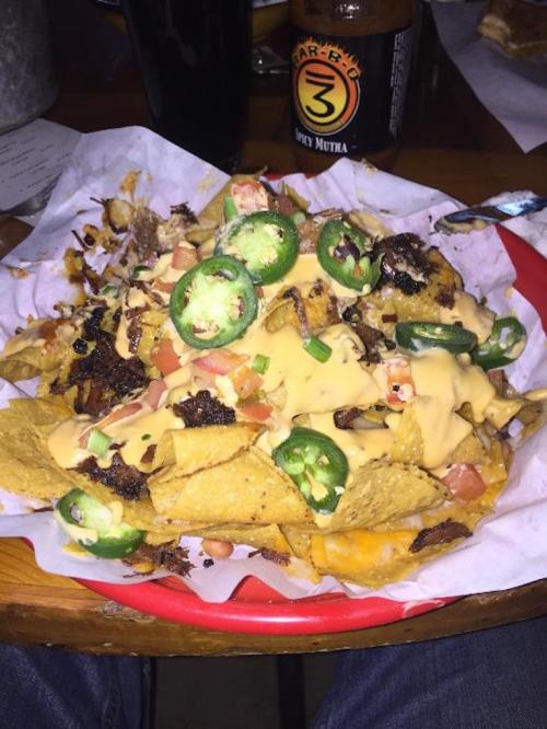 A plate of loaded nachos topped with cheese, jalapeños, diced tomatoes, and crispy bits, served with a drink in the background.
