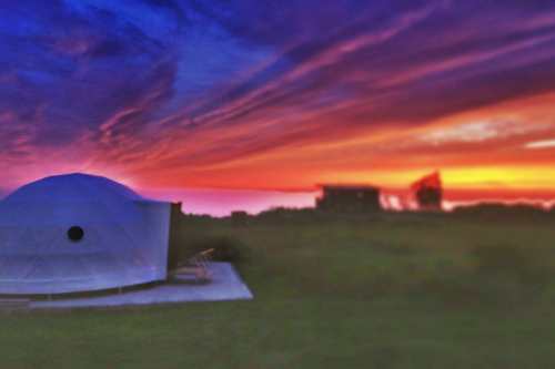 A geodesic dome structure against a vibrant sunset with colorful clouds and a distant building in the background.