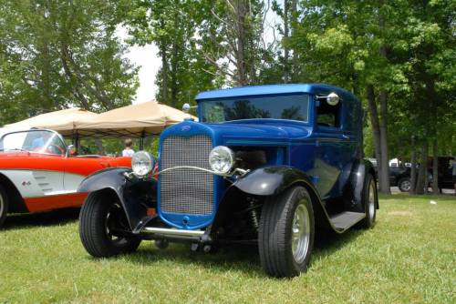 A classic blue vintage car parked on grass, surrounded by trees and another car in the background.
