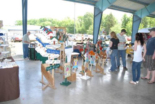 A craft fair scene with vendors displaying handmade items and visitors browsing under a large pavilion.