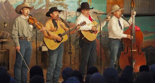 Four musicians in cowboy attire perform on stage with guitars, a fiddle, and a double bass, set against a scenic backdrop.