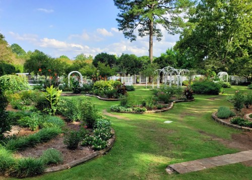 A lush garden with winding paths, colorful flowers, and white arbors under a blue sky with scattered clouds.