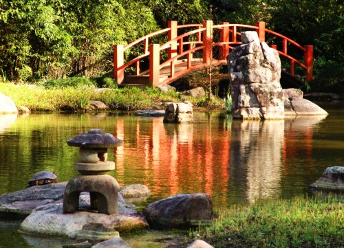 A serene garden scene featuring a red bridge, stone lantern, and rocks reflected in calm water surrounded by greenery.