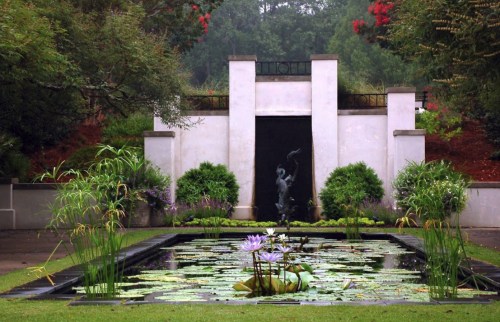 A serene garden with a pond filled with lily pads, a statue, and lush greenery in the background.