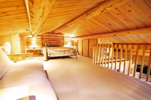 Cozy wooden loft with a bed, sofa, and warm lighting, featuring a sloped ceiling and wooden beams.