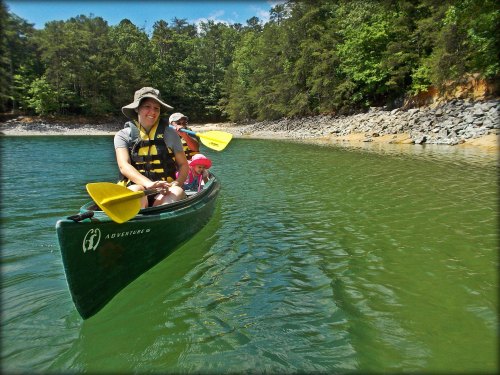 Two people in a canoe on a calm lake, surrounded by trees and rocky shoreline, enjoying a sunny day.
