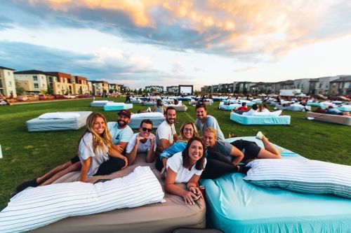 A group of nine friends lounging on beds outdoors, enjoying a movie night under a colorful sunset sky.
