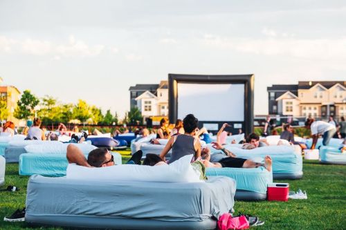 People lounging on inflatable couches in a grassy area, watching a movie on a large outdoor screen.