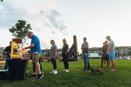 A group of people stands in line for popcorn at an outdoor event, with a dog and inflatable structure in the background.