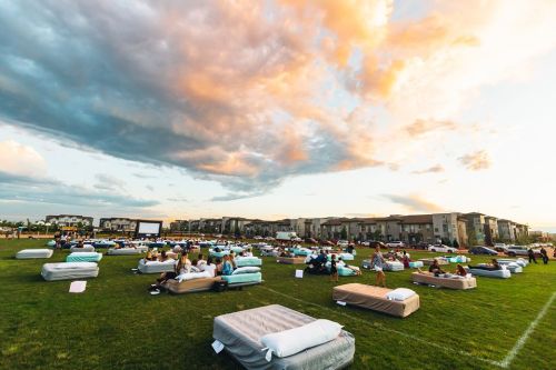 A large outdoor movie screening with people lounging on mattresses under a colorful sunset sky.