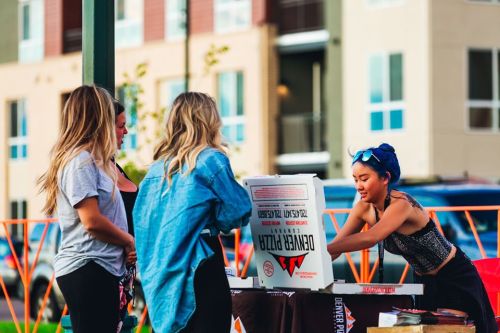 A group of four women gather around a pizza box at an outdoor event, smiling and chatting.